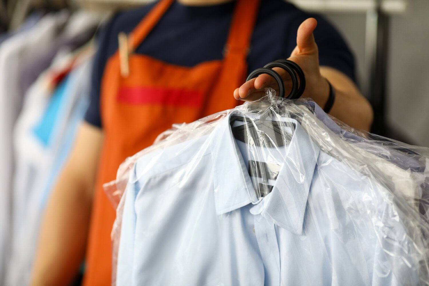 A person in an orange apron holds a freshly cleaned and pressed shirt covered in plastic at J Fields Cleaners, showcasing their quality dry cleaning and laundry service.