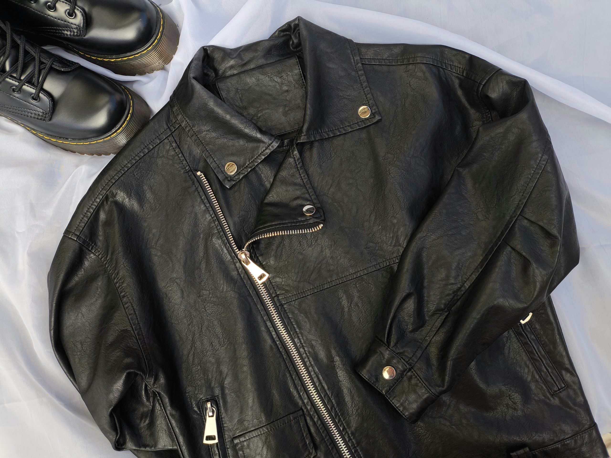 On a light gray background, a black leather jacket with an asymmetrical zipper and snap buttons sits next to lace-up boots, reflecting the high standards synonymous with Rancho Santa Margarita Suede & Leather Cleaning.