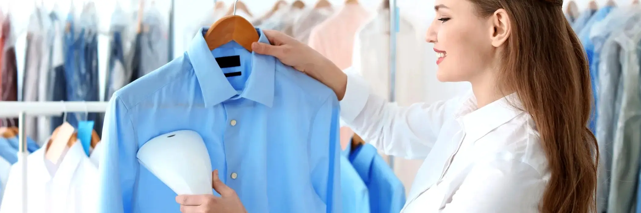 A woman uses a handheld garment steamer to remove wrinkles from a light blue dress shirt on a hanger, showcasing the quality care provided by J Fields Cleaners in a clothing store or laundry room.
