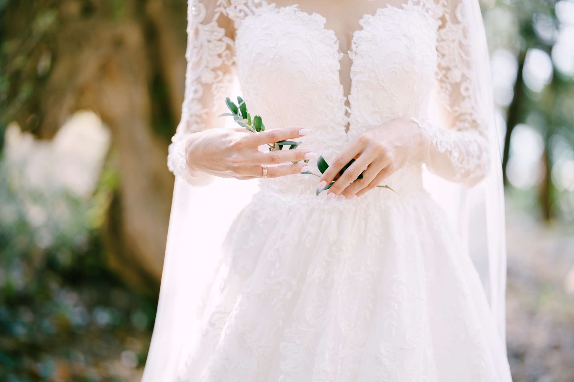 Bride in a lace wedding dress, standing gracefully with small green leaves in her hand against the blurred outdoor backdrop—perfect for a day that calls for special moments and memories worth preserving at Rancho Santa Margarita.