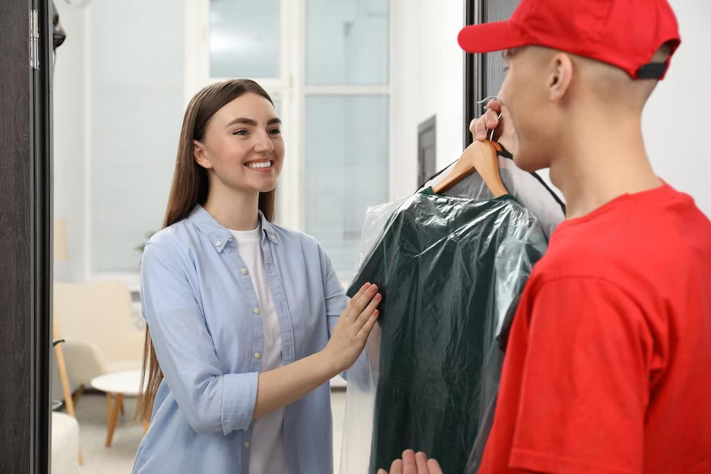A woman in a blue shirt receives a garment in plastic wrap on a hanger from a delivery person wearing a red cap and shirt, in a room with a beige chair and large windows.