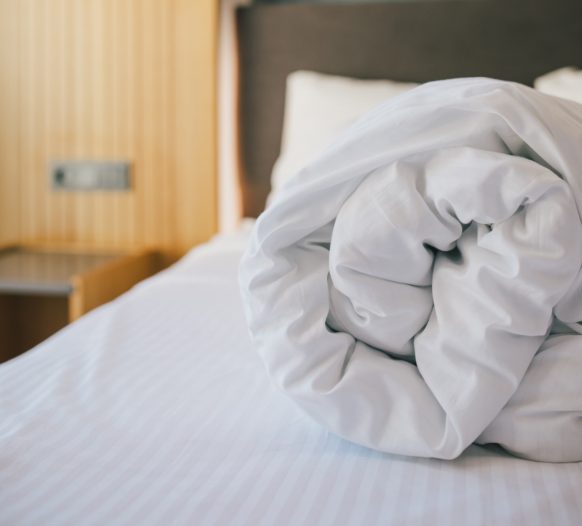 A neatly rolled white duvet lies on a white-striped bed in a warmly lit room, with a wooden nightstand and headboard in the background.