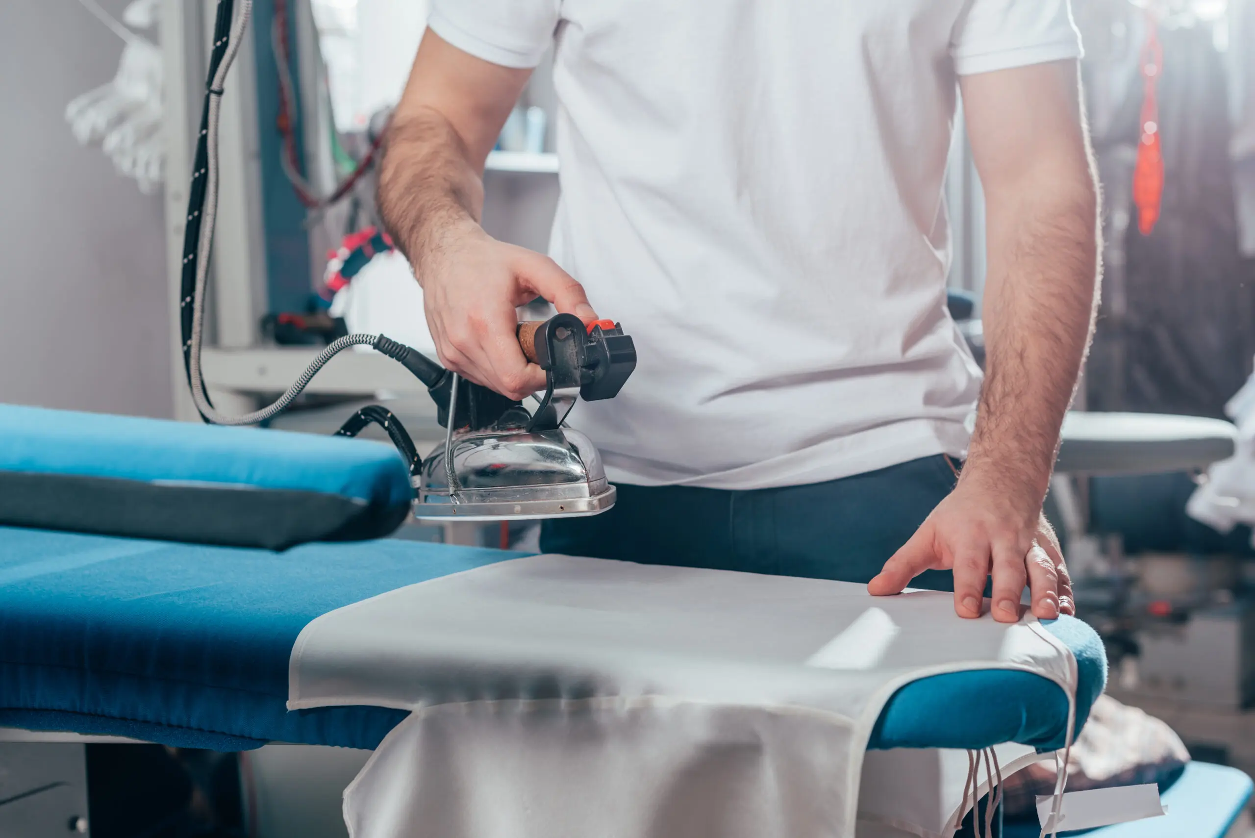 Person ironing a white cloth on a blue ironing board with a steam iron, in a well-lit room.