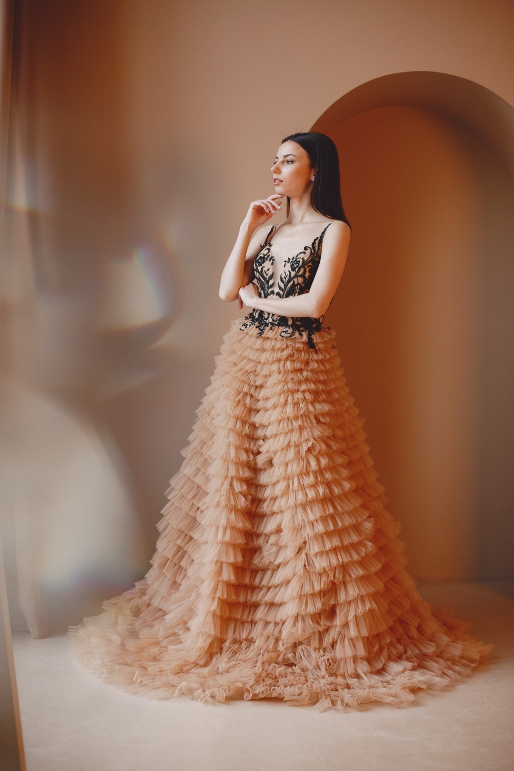 A woman stands thoughtfully by a window, wearing a flowing beige gown with a black lace bodice and layered ruffles.