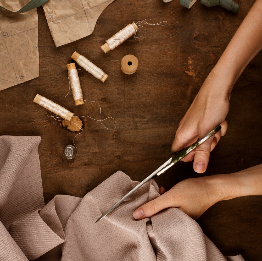 Hands cutting fabric with scissors on a wooden table, surrounded by spools of thread, a thimble, and sewing patterns.