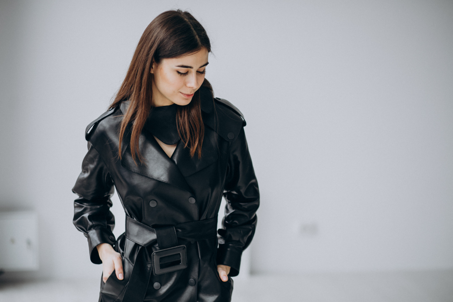 A person wearing a black leather trench coat with a wide belt stands against a plain light background, looking down with hands in pockets.