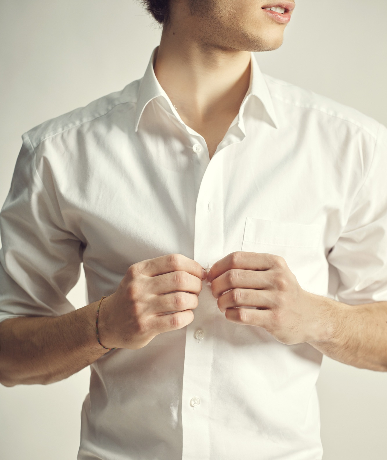 A person buttons a white dress shirt, with their head slightly turned, in a neutral, indoor setting.