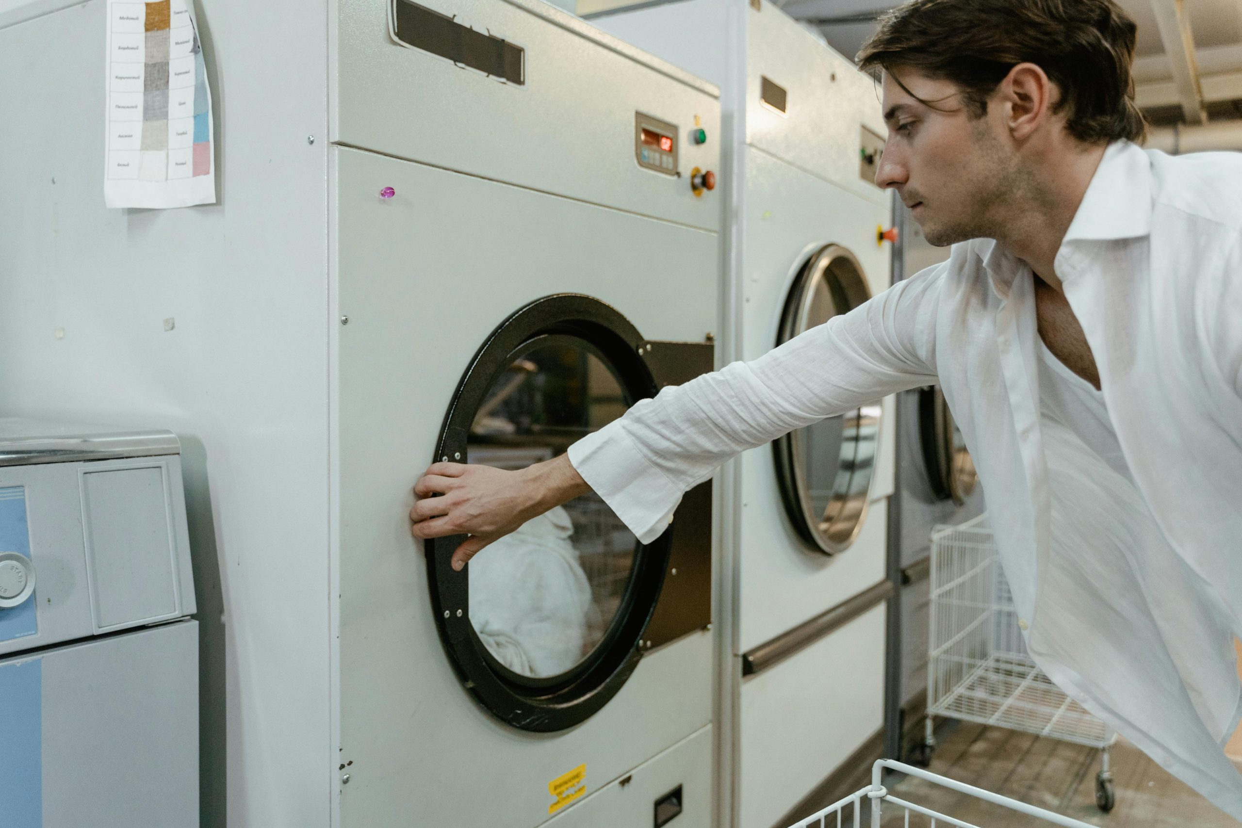 A person in a white shirt is using a laundromat dryer, placing their hand on the door.