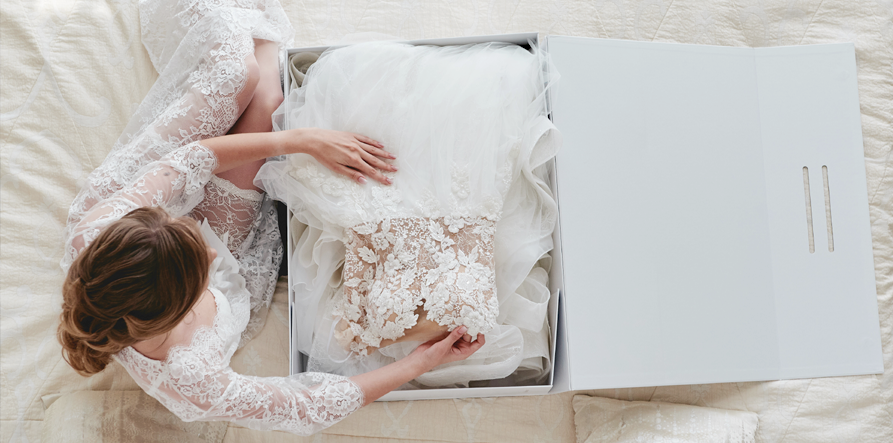 A woman in a lace robe unboxes a white wedding dress with floral lace details, placed in a white box on a bed.