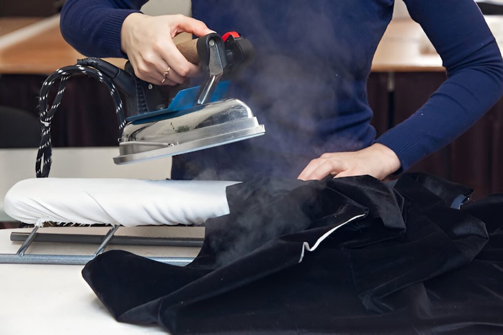 Person using a steam iron on a black velvet garment placed on an ironing board.
