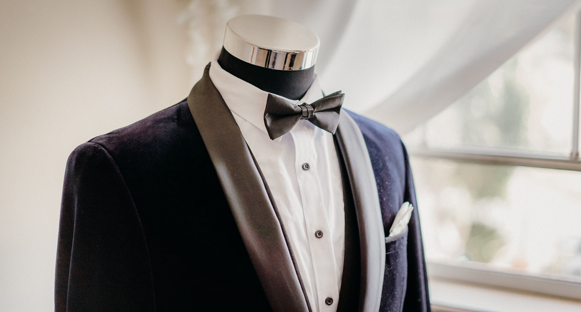 A tailor's mannequin displays a tuxedo with a black bow tie and a white dress shirt beside a window with sheer curtains.