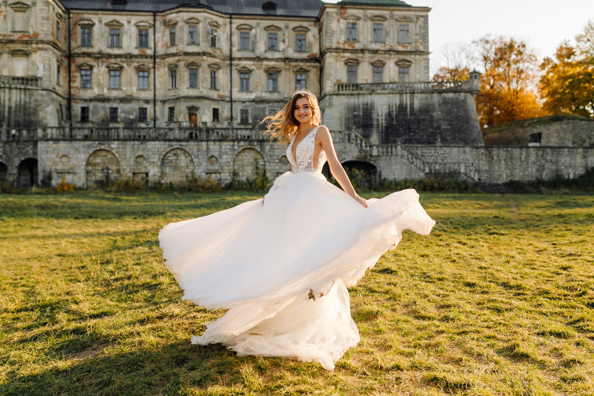 Bride in a flowing white dress twirls on grassy field with a historic stone building in the background.